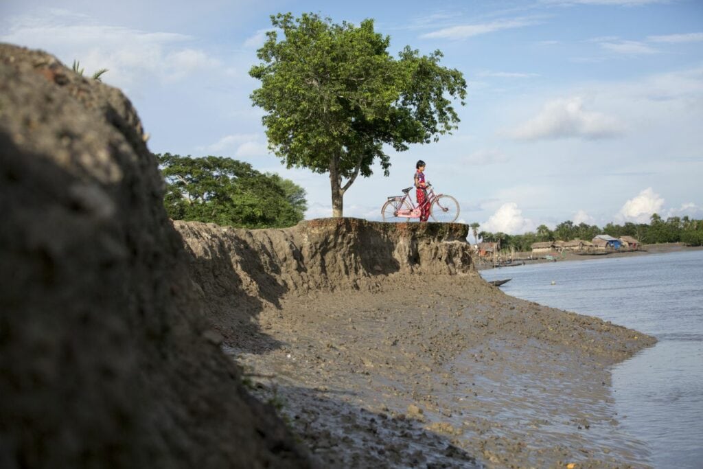 A coastline is affected by erosion. A boy stands with his bicycle at the edge.