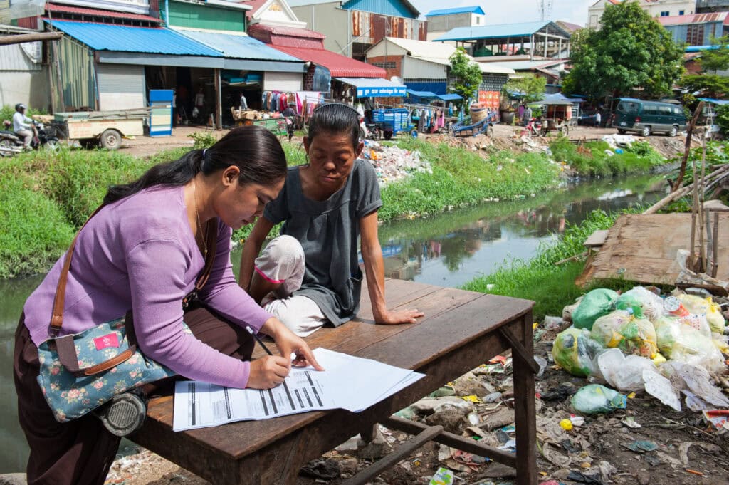 A woman is filling out a questionnaire, leaning over a table. Next to her, another person is sitting at the table, watching her fill it out.